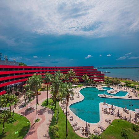 Royal Tulip Brasilia Alvorada Hotel Екстериор снимка The photo shows a modern hotel building with a striking red exterior, surrounded by lush greenery. In the foreground, there is a large, creatively shaped swimming pool, with lounge chairs arranged around it. The backdrop features a body of water, pos