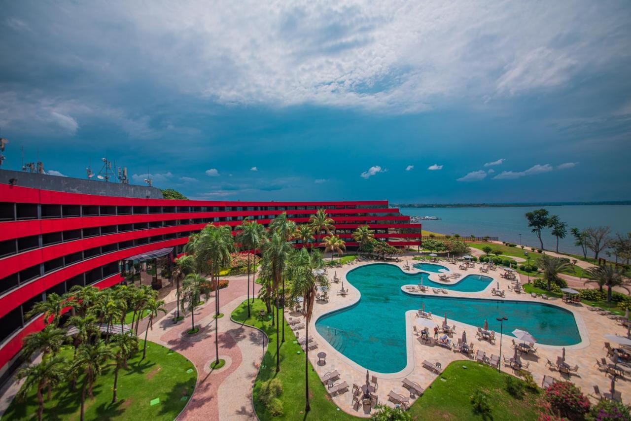 Royal Tulip Brasilia Alvorada Hotel Екстериор снимка The photo shows a modern hotel building with a striking red exterior, surrounded by lush greenery. In the foreground, there is a large, creatively shaped swimming pool, with lounge chairs arranged around it. The backdrop features a body of water, pos