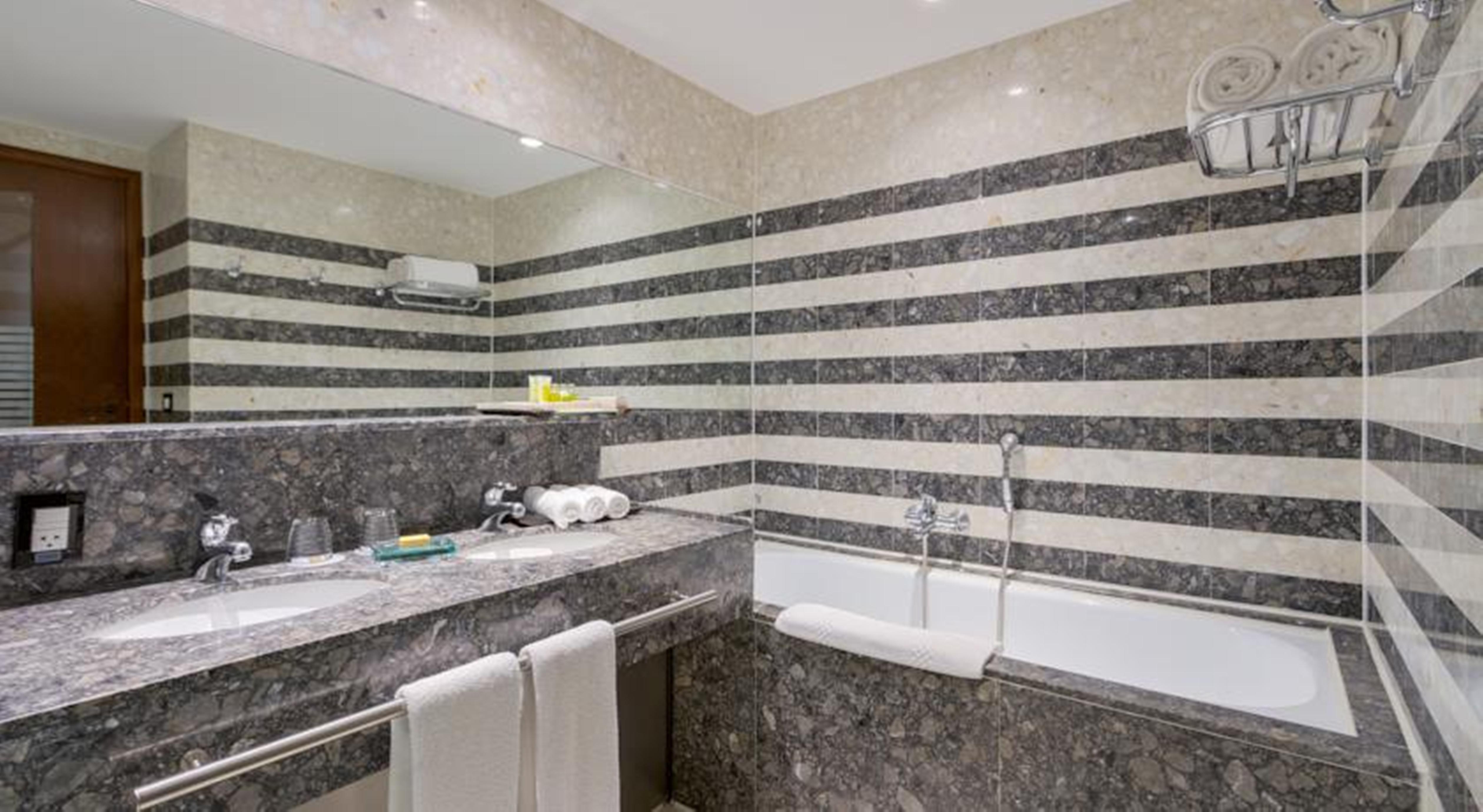 Royal Tulip Brasilia Alvorada Hotel Екстериор снимка The photo shows a modern bathroom featuring a combination of marble and stone finishes. The walls are decorated with horizontal stripes in various shades of gray and cream. There is a bathtub with a white exterior, and a showerhead is mounted on the 