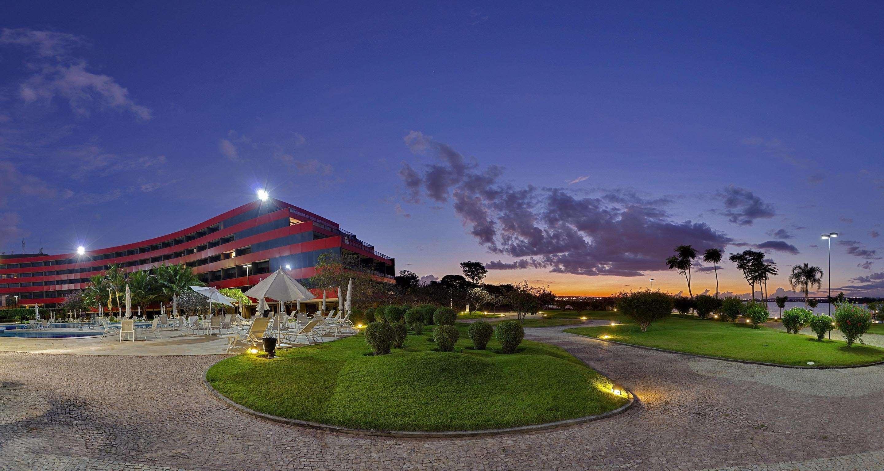 Royal Tulip Brasilia Alvorada Hotel Екстериор снимка The photo depicts a modern building, possibly a hotel or resort, with a striking red exterior. It is set in a landscaped area that features well-maintained greenery and ornamental plants. In the foreground, there are lounge areas with umbrellas, sugg