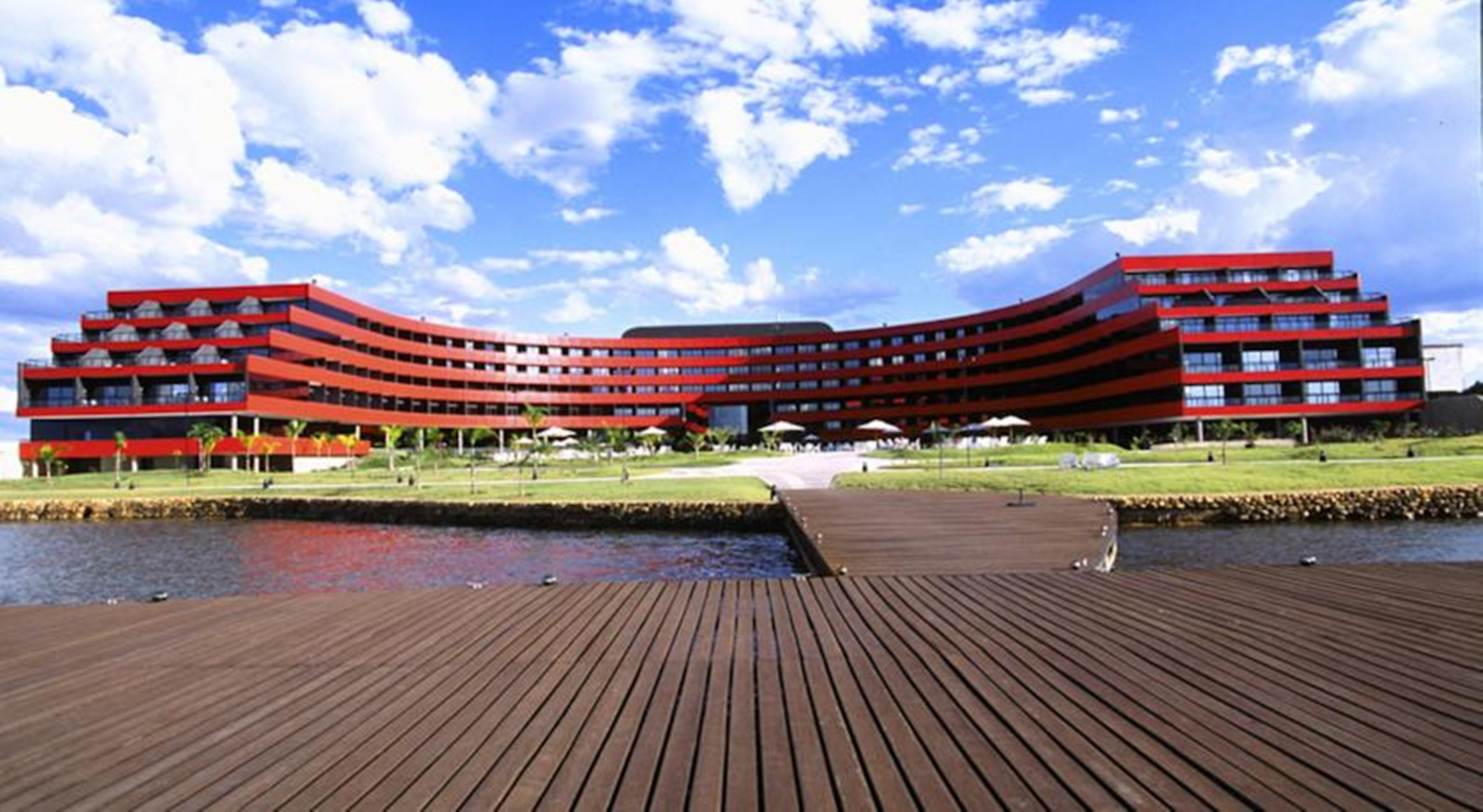 Royal Tulip Brasilia Alvorada Hotel Екстериор снимка The image shows a modern, curved building with a striking red facade, situated near a body of water. The architecture features multiple levels with large windows that create a sleek and contemporary appearance. In front of the building, there is a wo