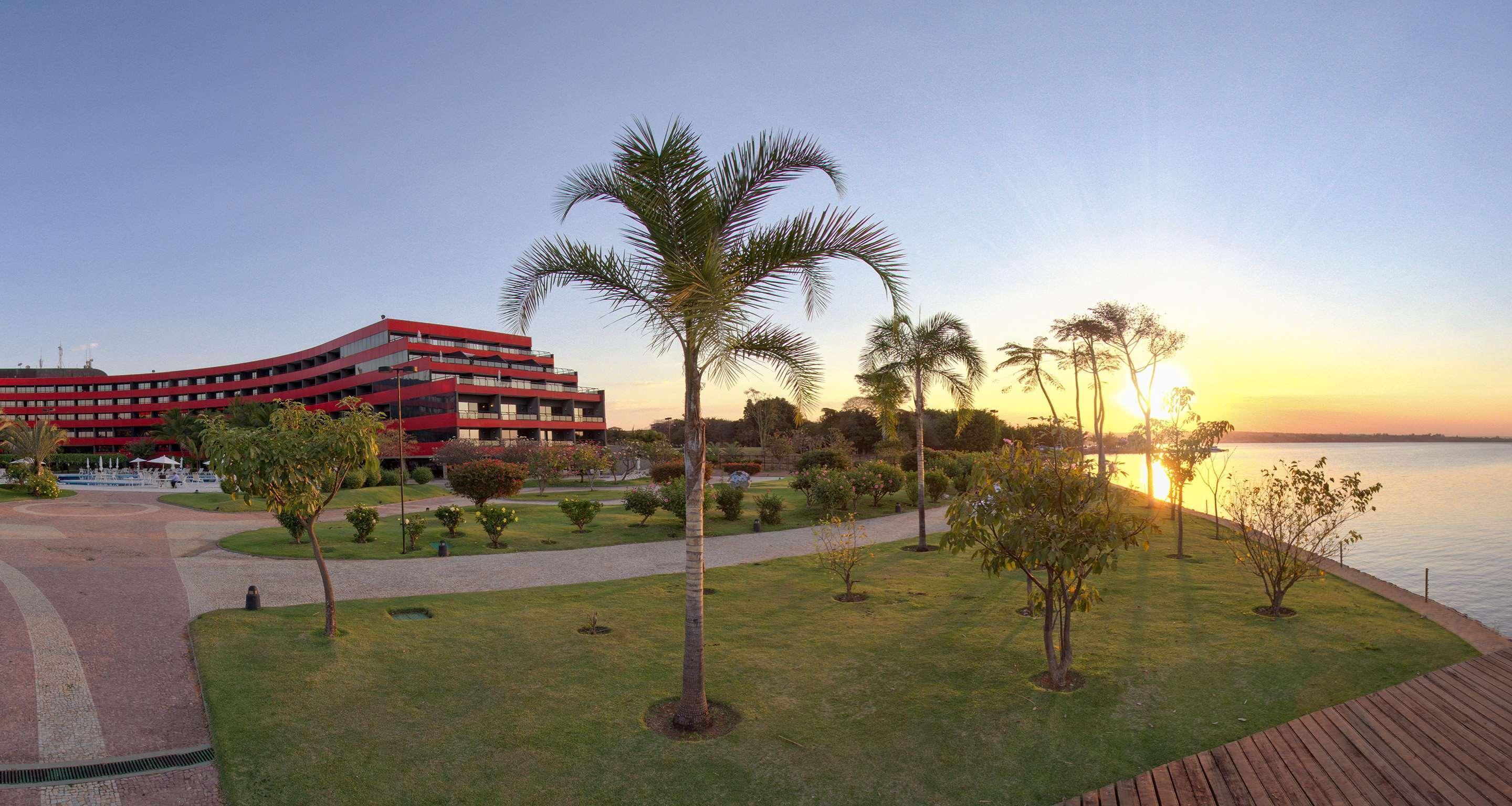 Royal Tulip Brasilia Alvorada Hotel Екстериор снимка The photo depicts a scenic view featuring a red multi-story building alongside a serene waterfront. In the foreground, there are several palm trees and lush green lawns, creating a tropical atmosphere. The sun is setting in the background, casting wa