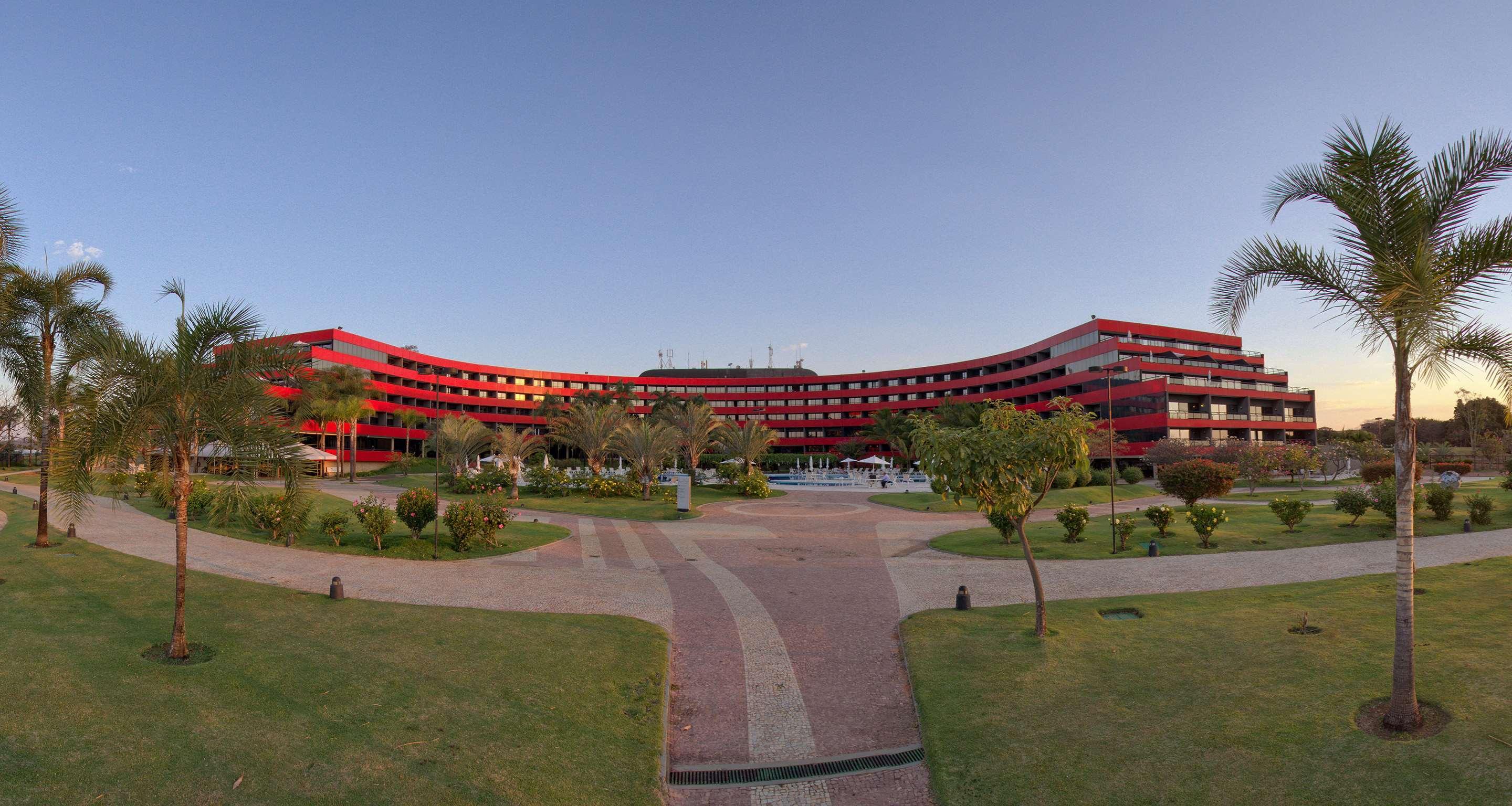 Royal Tulip Brasilia Alvorada Hotel Екстериор снимка The photo shows a large, curved building with a vibrant red exterior. It appears to be a hotel or resort, surrounded by well-maintained gardens and green spaces. The landscape includes manicured lawns, small trees, and pathways, suggesting a pleasant