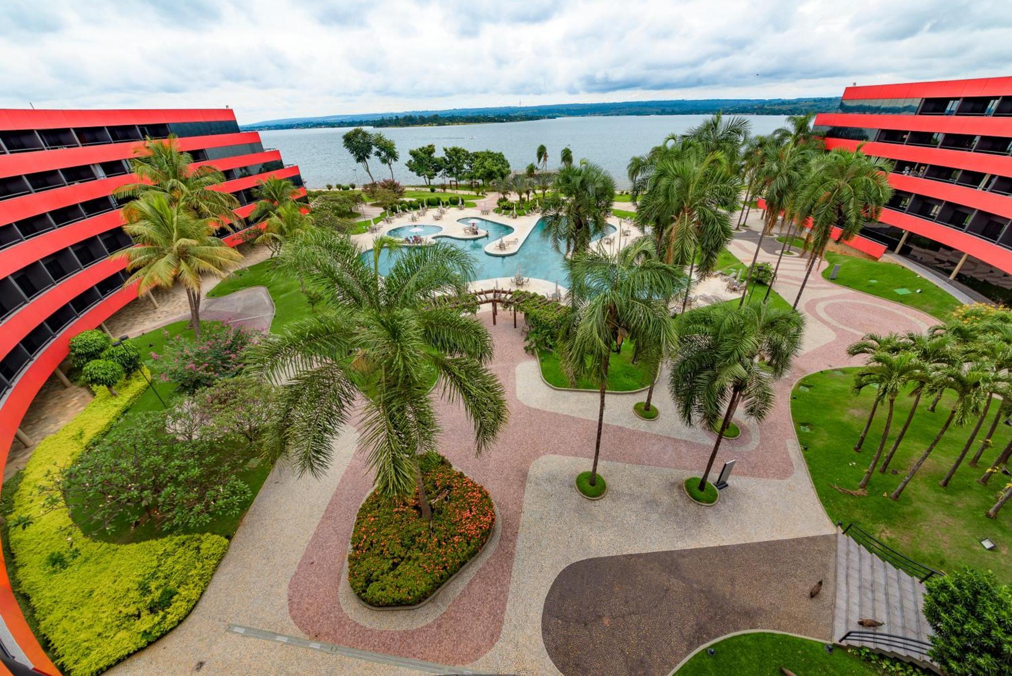 Royal Tulip Brasilia Alvorada Hotel Екстериор снимка The photo shows a scenic view of a courtyard area in a residential complex. There are multiple red buildings surrounding a pool area, which is surrounded by palm trees and landscaped gardens. The pool features a curvy design, and there are pathways m